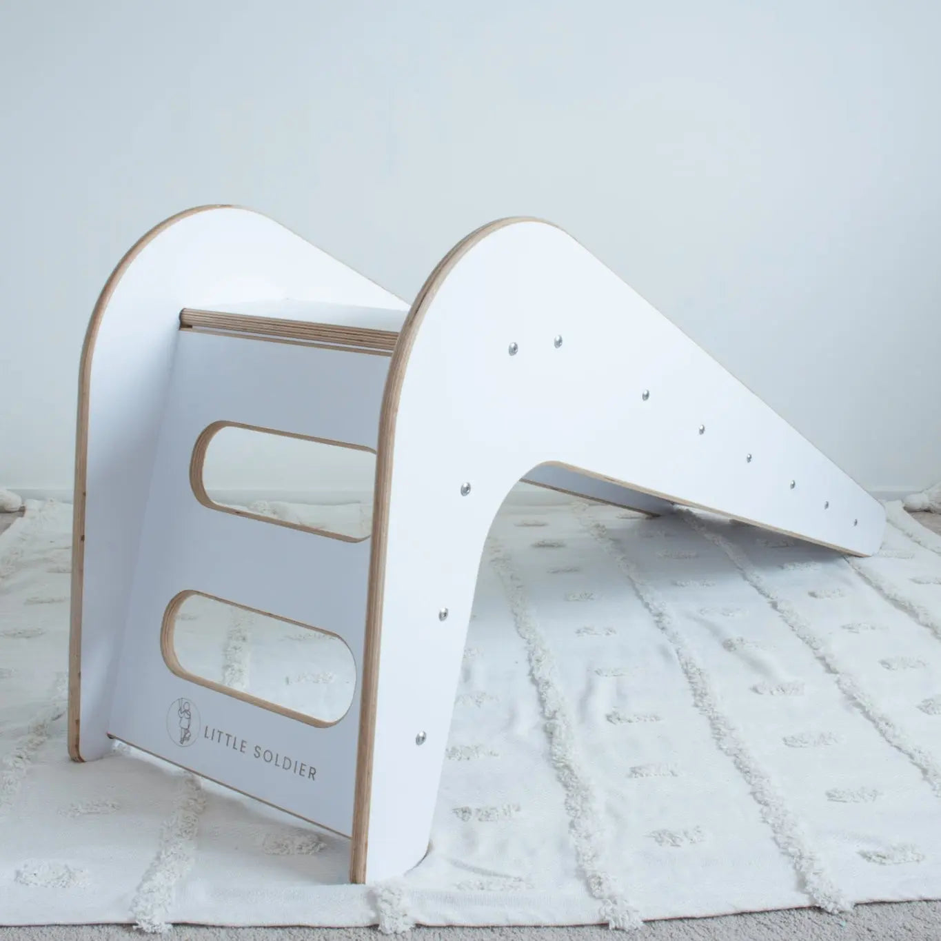 A wooden, white slide shown from its back-left side sitting on top of a white tufted rug. The image shows the steps of the slide and the Little Soldier logo