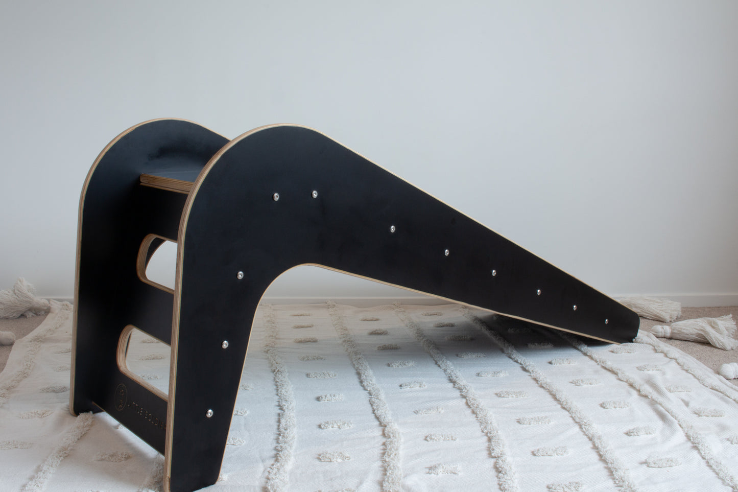 A black wooden slide shown from its back-left side sitting on top of a white tufted rug. The image shows the steps of the slide.