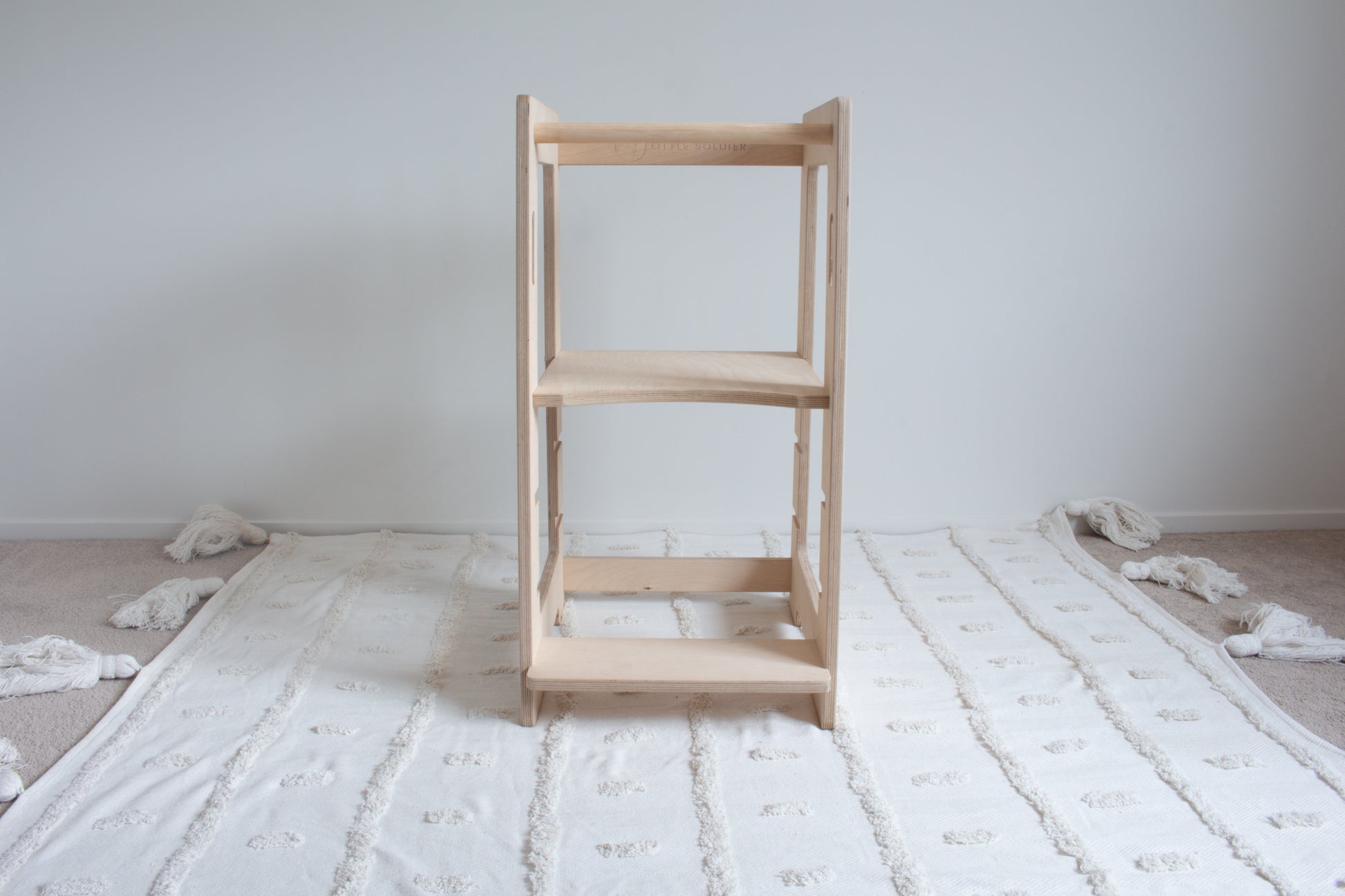 A natural wooden kitchen helper viewed front on sitting on top of a tufted rug. The adjustable platform is set to the highest setting.