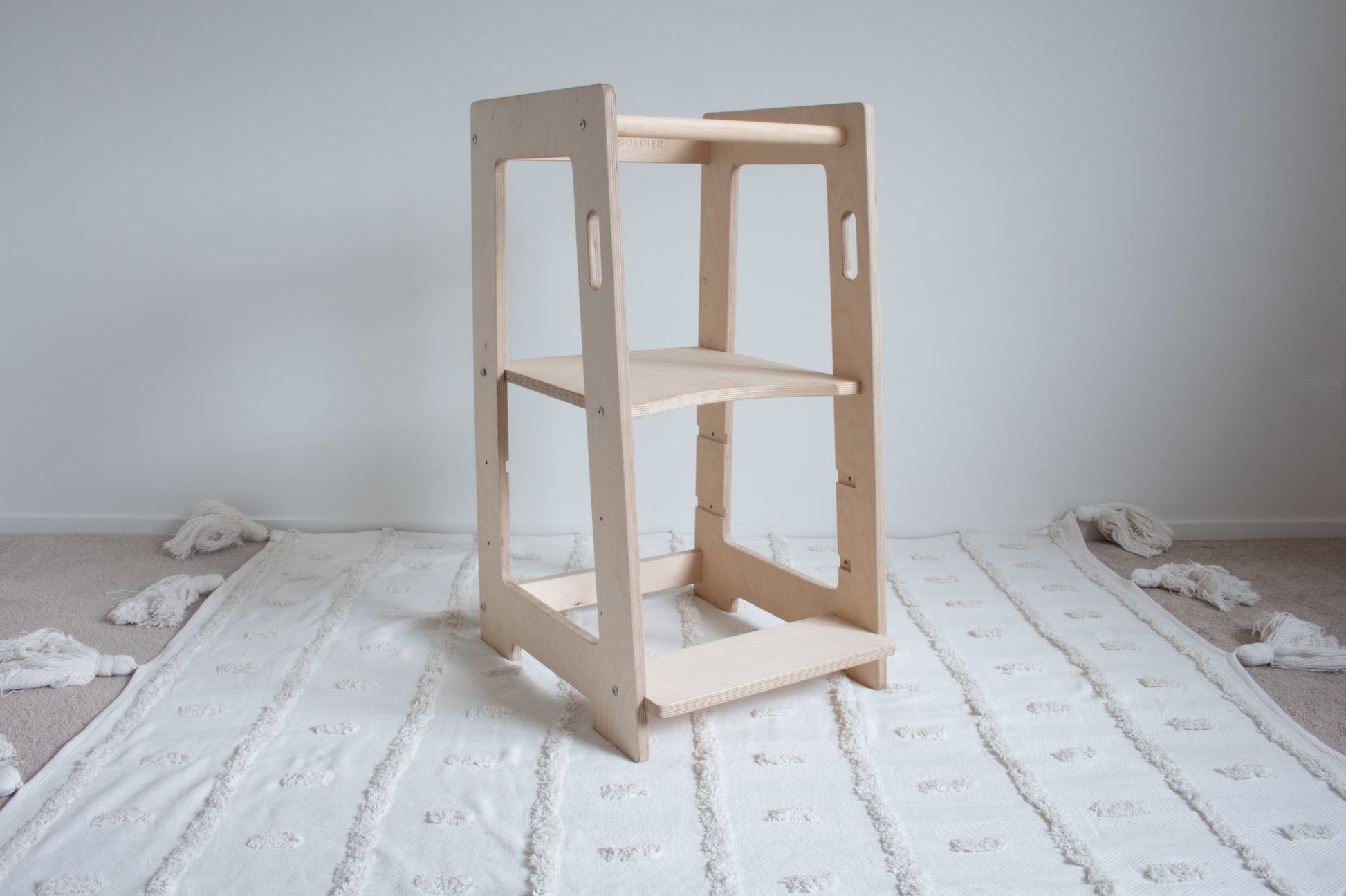 A natural wooden learning tower viewed from the left front side sitting on top of a tufted rug. The adjustable platform is set to the highest setting.