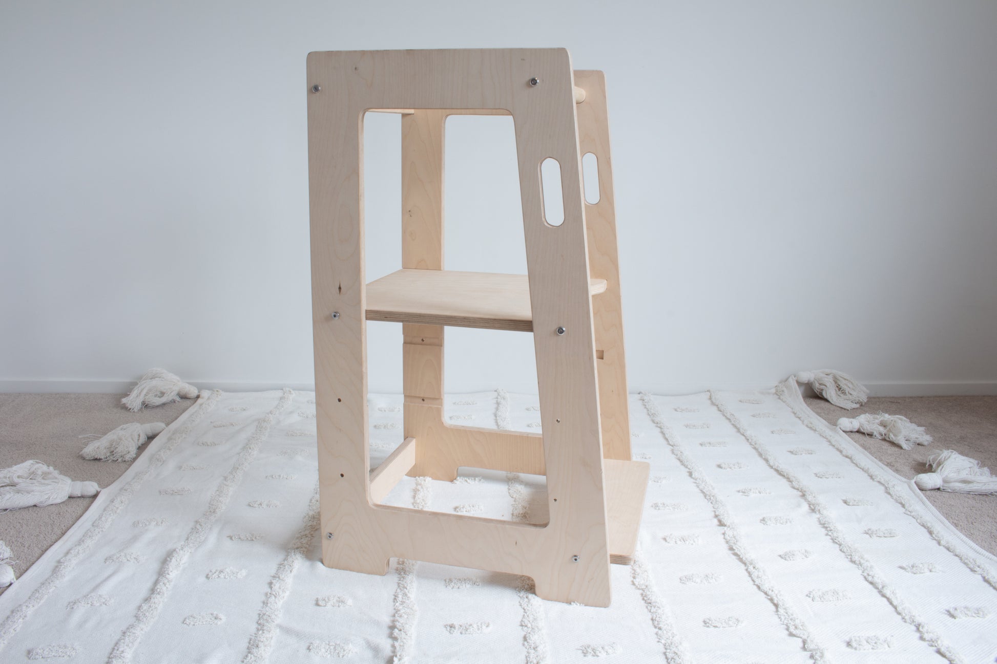 A natural wooden learning tower viewed from the left side sitting on top of a tufted rug. The adjustable platform is set to the highest setting.