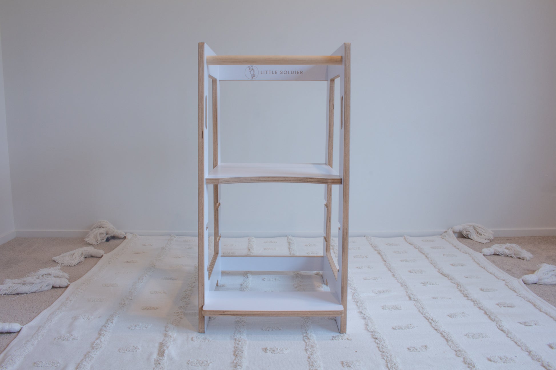 A white learning tower viewed front-on sitting on top of a tufted rug. The adjustable platform is set to the highest setting. The Little Soldier logo can be seen.